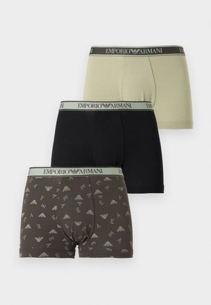 Mężczyzna bez koszulki stojący boso, ubrany w ciemne bokserki w wzory z logiem Emporio Armani na gumce, na jednolitym tle.