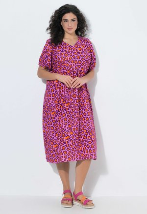 Kvinde med krøllet hår iført en lilla og orange leopardprintet midikjole og pink-orange sandaler, stående mod en hvid baggrund.