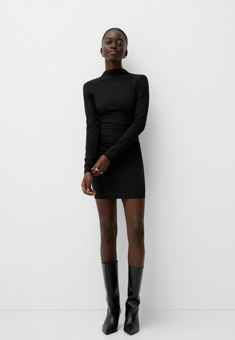 Vestido negro de manga larga con diseño ajustado y cuello alto, combinado con botas negras hasta la rodilla, que destacan una textura suave.