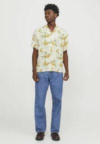Camisa de manga corta y botones en amarillo pálido con un patrón de frutas naranjas y hojas verdes, combinada con unos jeans azules y zapatos marrones oscuros.