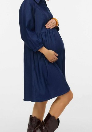 Gravid kvinde iført en mørkeblå knap-op kjole, brune ankelstøvler og et rav-armbånd, som holder om sin mave, stående sideværts.