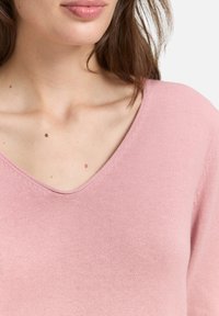 Pull-over V en cachemire rose clair avec une texture douce et lisse ; il présente un col côtelé et des épaules légèrement tombantes.