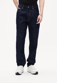 Jeans de mezclilla azul oscuro con un corte relajado, que presentan costuras naranjas, cinco bolsillos y una cremallera, combinados con zapatillas blancas.