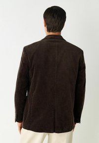 Bruine corduroy blazer met een gestructureerd ontwerp, voorzien van een met leren kraag en een enkele split aan de achterkant. Vloeiende textuur met zichtbare ribbels.