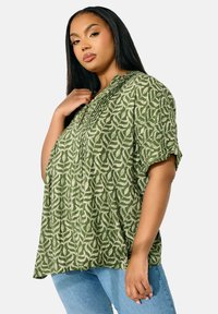 Grön mönstrad blus med korta ärmar och knappdetaljer. Har en strukturerad design av vita bladformer mot en enfärgad bakgrund.