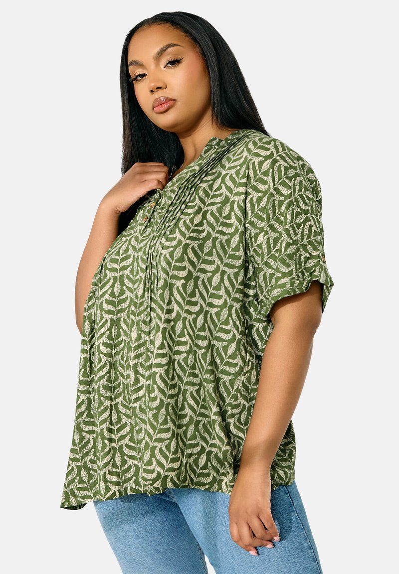 Grön mönstrad blus med korta ärmar och knappdetaljer. Har en strukturerad design av vita bladformer mot en enfärgad bakgrund.