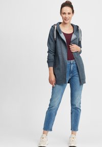 Mørkeblå zip-up hettegenser med lyse grå detaljer, snor og lommer, kombinert med en kastanjefarget topp og lyseblå jeans. Hvite joggesko.
