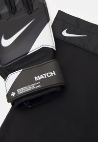 Guantes de portero negros con palma texturada, acentos en blanco y gris, y una correa elástica negra en la muñeca etiquetada "MATCH."