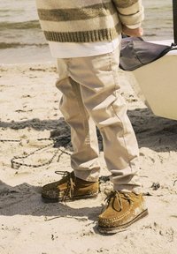 Smeđe suède cipele s vezanjem i dekorativnim šavovima, u paru s svijetlosmešnim hlačama, stoje na pješčanoj plaži blizu vode.