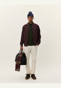 Jachetă bomber bordeaux peste o cămașă butonată verde închis, pantaloni crem, pantofi închiși la culoare, căciulă bleumarin, ținând o geantă neagră cu o eșarfă cu carouri.