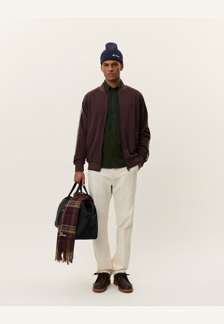 Jachetă bomber bordeaux peste o cămașă butonată verde închis, pantaloni crem, pantofi închiși la culoare, căciulă bleumarin, ținând o geantă neagră cu o eșarfă cu carouri.