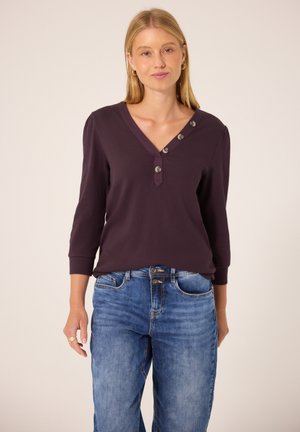 Femme blonde portant un haut à manches longues violet foncé avec des boutons inclinés et un jean bleu, debout devant un fond uni.