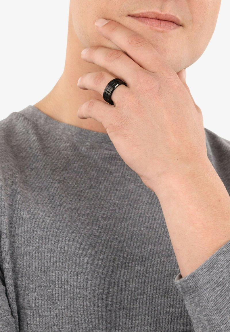 Anillo negro con un diseño texturizado que presenta secciones alternas suaves y estriadas, llevado en una mano sobre un fondo de suéter gris.
