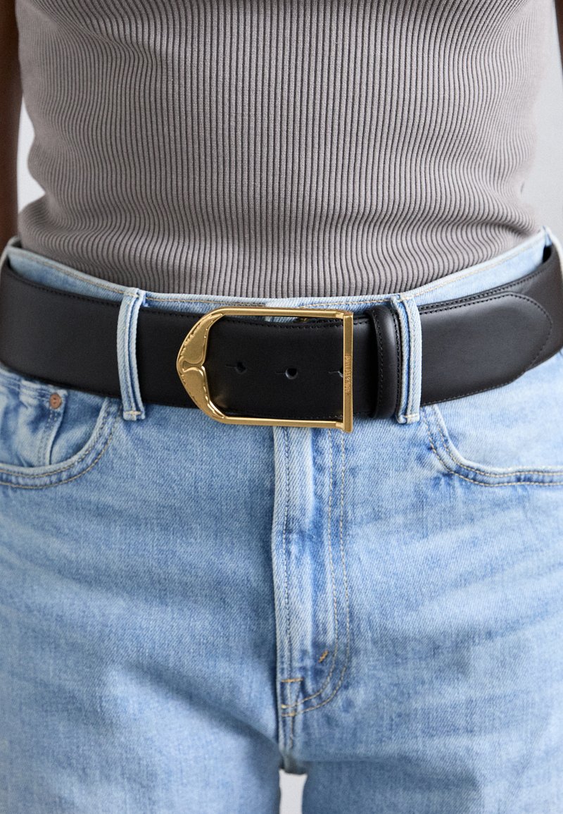 Black leather belt with a gold metal buckle featuring a curved design, worn over light blue denim jeans.