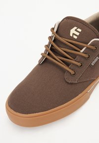 Brown canvas shoe with brown laces, rubber sole, and a textured upper. Features a logo on the tongue and stitching along the edges.