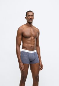Boxershorts med et navyblåt teksturmønster, der har en hvid linning mærket "TOMMY HILFIGER." Stoffet virker strækbart og tætsiddende.