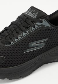 Black athletic shoe with a textured mesh upper, featuring a cushioned sole, and reflective accents. Includes a logo on the side.