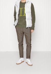 Personne portant une veste matelassée blanche, un pull vert avec des lettres jaunes, un pantalon à carreaux marron et des baskets blanches, debout devant un fond uni.