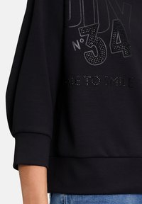 Sweat-shirt noir avec manches bouffantes, comportant des lettres en relief en argent et des strass inscrivant "No 34" et "TIME TO SMILE."
