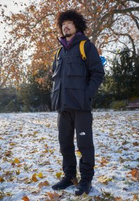Jachetă neagră rezistentă la intemperii, cu multiple buzunare, purtată împreună cu pantaloni negri și cizme. Rucsac galben cu curele albastre. Pământ acoperit cu zăpadă și frunze.