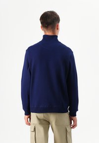 Una persona sta in piedi con le spalle rivolte verso la telecamera, indossando un maglione a collo alto blu navy e pantaloni cargo beige chiaro su uno sfondo bianco.