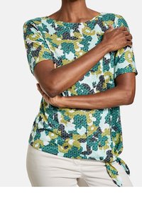 Top de manga corta multicolor con un patrón floral texturizado en verde, negro y blanco; presenta un detalle de lazo en el lateral y un cuello redondo.