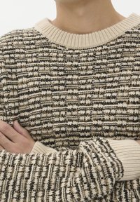 Pleteno pulover z bež in črno teksturo, z rebrastim izrezom in manšetami. Tkanina se zdi topla in udobna.