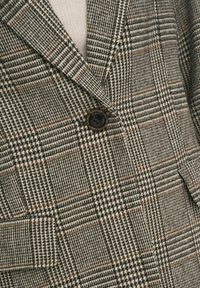 Veste à carreaux pied-de-poule marron, beige et noir avec un unique bouton foncé et des poches à rabat à l'avant.