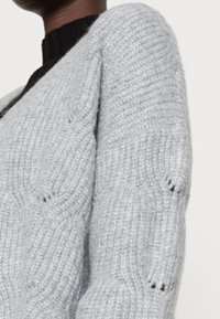 Grå stickad kofta med ribbat mönster och V-ringning, med synlig sömnad och en mjuk, fluffig look.