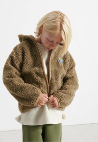 Bambino con capelli biondi che indossa una giacca di pile marrone sopra una maglietta crema e pantaloni verdi, che guarda in basso mentre chiude la cerniera della giacca.