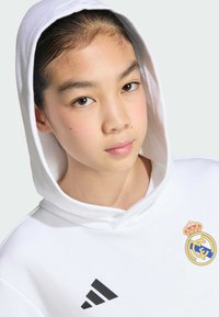 Sweat à capuche blanc avec une grande capuche, arborant le logo brodé du Real Madrid et des bandes noires Adidas. Texture de tissu lisse, design simple.