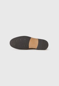 Suela de zapato de cuero marrón con una base de goma negra, que presenta seis tracciones circulares y una textura suave. Detalle de costura visible cerca del talón.