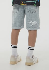 Shorts in denim con risvolti, in un blu chiaro, due tasche posteriori e testo in stile graffiti bianco sul lato. Abbinati a sneakers bianche e calzini a righe.