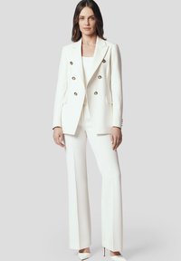 Biała marynarka o dopasowanym kroju z podwójnym zapięciem na złote guziki, zestawiona z białymi spodniami o szerokich nogawkach i pasującymi szpilkami z wąskim noskiem.