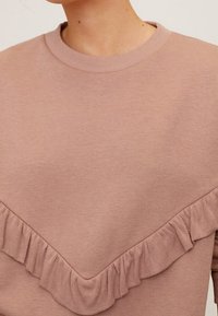 Jasnoróżowa bluza wykonana z miękkiego materiału, z fakturowanym detalem w postaci falbanki wzdłuż dekoltu w kształcie litery V i żebrowanymi brzegami.