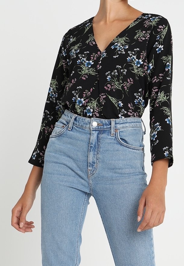 Personne portant un chemisier noir à manches longues à motifs floraux, rentré dans un jean bleu clair taille haute avec poches avant et fermeture par boutons.