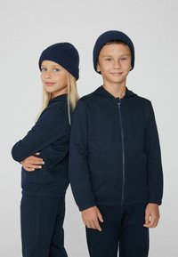 Dwoje dzieci w dopasowanych granatowych bluzach z zamek i prążkowanych czapkach. Materiał wydaje się miękki o gładkiej fakturze i luźnym kroju.