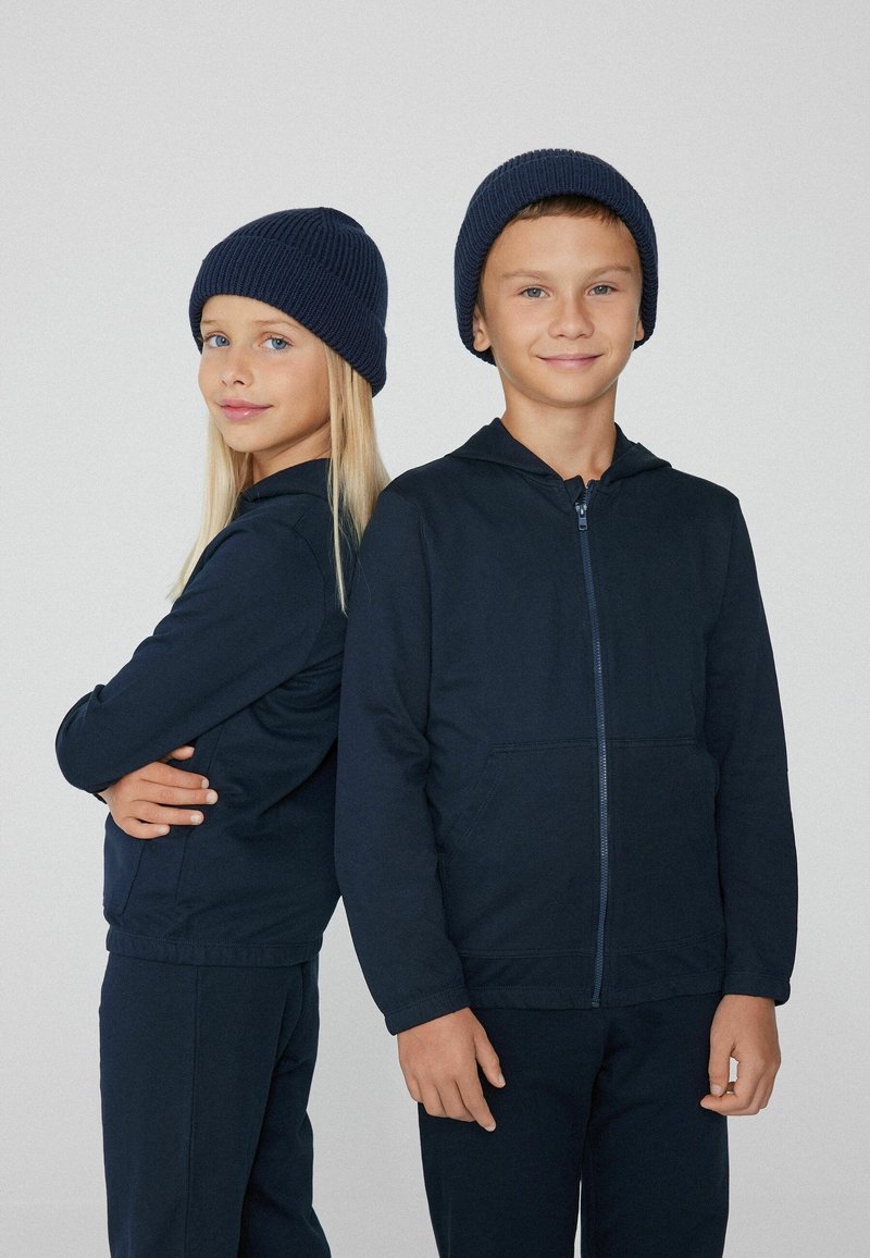 Dwoje dzieci w dopasowanych granatowych bluzach z zamek i prążkowanych czapkach. Materiał wydaje się miękki o gładkiej fakturze i luźnym kroju.