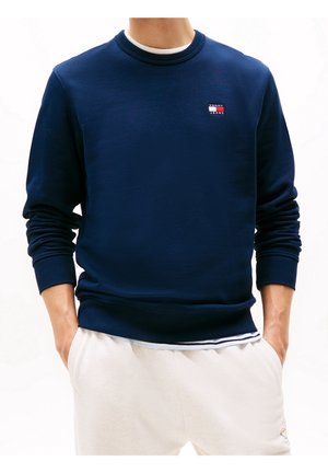 Mężczyzna ubrany w granatową bluzę z logo Tommy Jeans na piersi i białe spodnie, ręce w kieszeniach, na jednolitym białym tle.