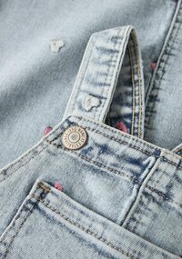 Šviesiai mėlynos spalvos džinsiniai kombinezonai su išblukusia tekstūra, širdies rašto siuvinėjimu ir metaline aparatūra ant petnešos bei kišenės srityje.