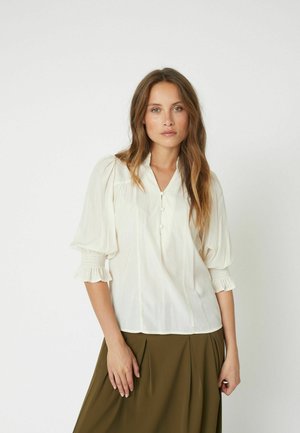 White, lightweight blouse with a V-neck, short puff sleeves, and buttoned placket. Paired with a green skirt, featuring gentle pleats.