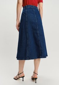 Denim midi sukňa v tmavomodrej farbe s bezšvovým dizajnom, ukončeným lemom a vysokým pásom. Skombinovaná s čiernymi sandálmi na podpätku.