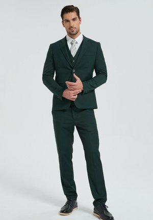 Homme debout portant un costume vert foncé avec gilet, cravate claire à motifs, chemise blanche et chaussures habillées noires sur un fond uni.