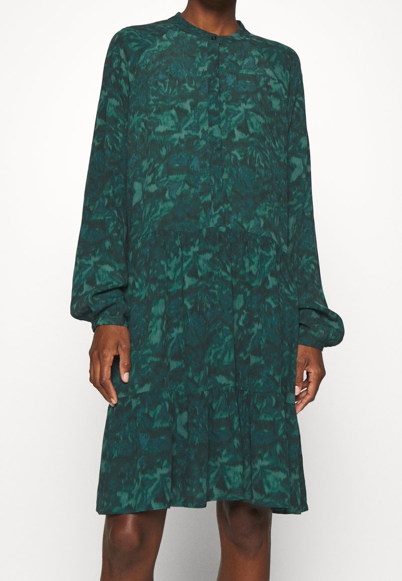 Robe longue à manches longues à motif vert foncé, col boutonné, coupe ample, longueur genou, portée par une personne à la peau foncée.