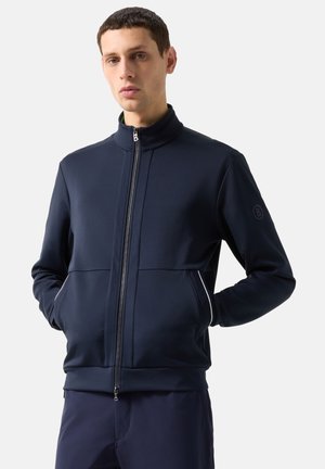 Mężczyzna ubrany w granatową kurtkę na zamek z wysokim kołnierzem, z rękami w kieszeniach, stojący na białym tle.