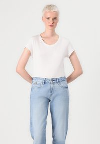 Witte korte mouwen t-shirt met een v-hals, gecombineerd met lichtblauwe high-waisted jeans. Model staat met de handen op de heupen.