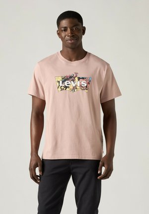 Junger Mann trägt ein hellrosa Levi's T-Shirt mit buntem Logo und schwarzen Hosen, steht vor einem schlichten hellen Hintergrund.