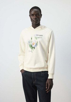 Hombre con sudadera con capucha color crema con estampado floral difuminado y texto "Hidden Nature Series 2026,", combinado con vaqueros azul oscuro, de pie frente a un fondo liso.