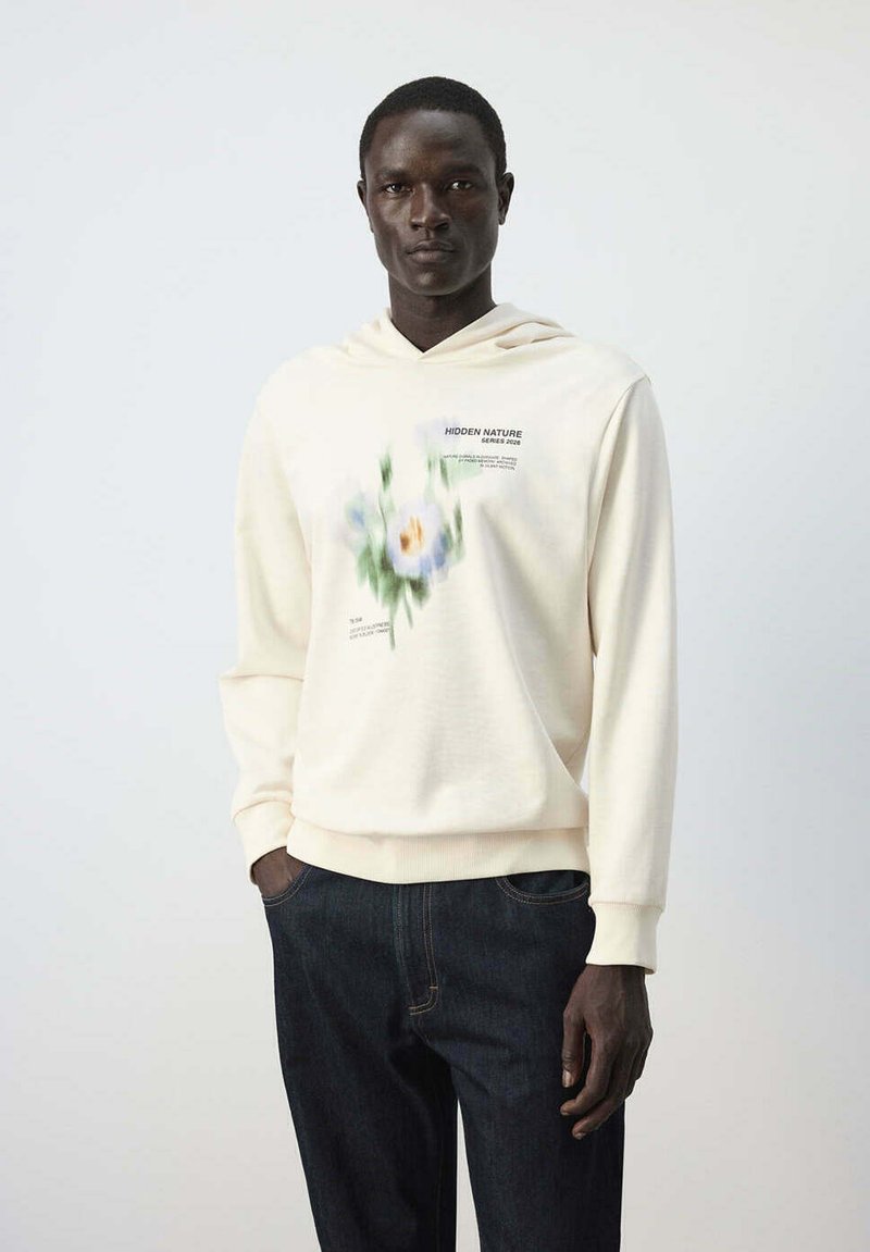 Homme portant un sweat à capuche crème avec imprimé floral flou et texte "Hidden Nature Series 2026,", associé à un jean bleu foncé, debout devant un fond uni.