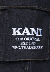 Mörk denimtyg med en broderad logotyp som säger "KANI THE ORIGINAL EST. 1989 REG. TRADEMARK" i vitt, accentuerad med gula stygn.